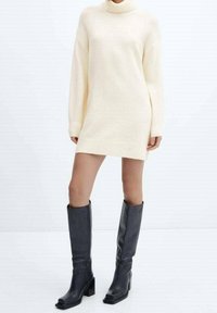 Žena v krémovém roláku a černých kozačkách na vysokém podpatku, stojící na bílé podlaze.