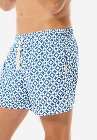 Badehose mit geometrischem Muster in Blau, Grün und Weiß, ausgestattet mit einem elastischen Bund und einer Kordel, aus leichtem Stoff gefertigt.