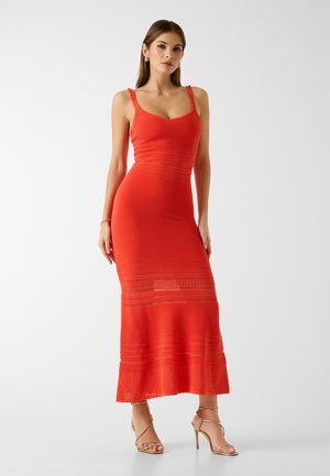 Kobieta w dopasowanej, bezrękawnikowej czerwonej sukni maxi z koronkowymi detalami, nosząca złote, wysokie sandały na paskach, stoi na białym tle.