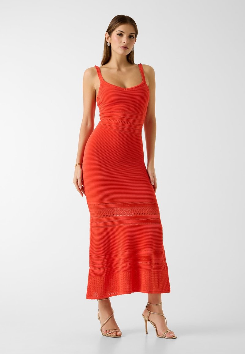 Kobieta w dopasowanej, bezrękawnikowej czerwonej sukni maxi z koronkowymi detalami, nosząca złote, wysokie sandały na paskach, stoi na białym tle.