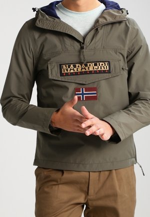 Osoba nosząca oliwkową kurtkę Napapijri z naszywką norweskiej flagi i logo na piersi, w połączeniu z brązowymi spodniami.