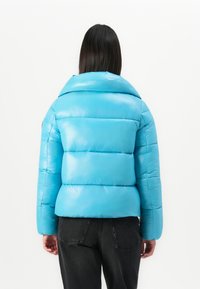 Persoon met lang donker haar, gekleed in een helderblauwe pufferjack en zwarte jeans, gezien van achteren tegen een witte achtergrond.