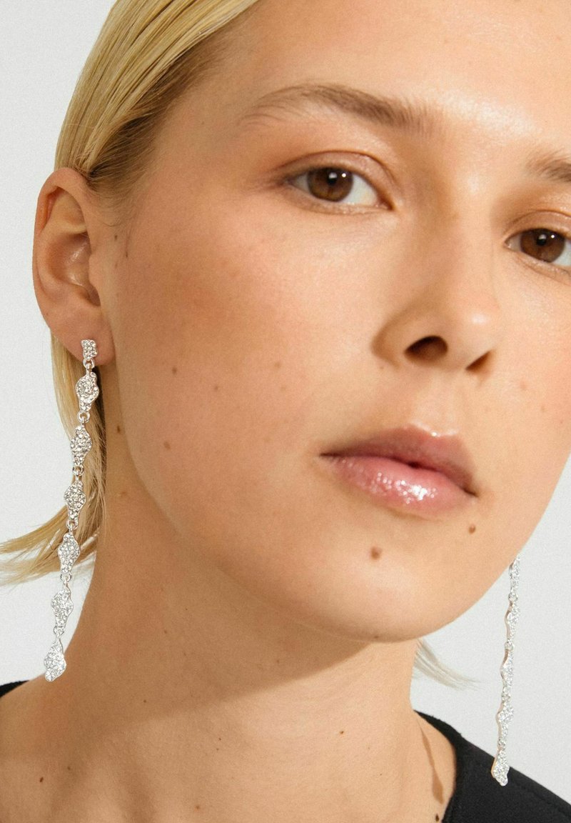 Mujer con cabello rubio corto, llevando pendientes largos de plata texturizados y labios rosas brillantes, contra un fondo liso claro.