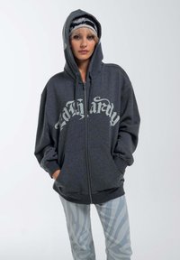 Gray zip-up hoodie with large white script logo on the front, featuring a spacious hood and front pockets, paired with light striped pants.