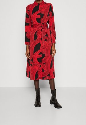 Czerwona i czarna sukienka z wirującym wzorem, długimi rękawami, kołnierzykiem i wiązanym w pasie. W połączeniu z czarnymi botkami za kostkę.