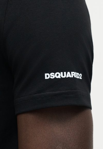 Μαύρο κοντομάνικο μπλουζάκι σε σκουρόχρωμο χέρι με λευκό λογότυπο "DSQUARED2" τυπωμένο κοντά στο τελείωμα του μανικιού.
