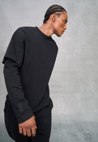 Schwarzes Baumwoll-T-Shirt, über einem Langarmhemd getragen, mit einem lockeren Schnitt und dezenten bestickten Details an den Ärmeln.