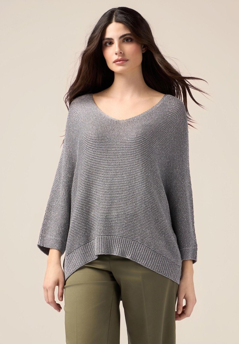 Grauer gestrickter Pullover mit entspanntem Schnitt, geripptem Saum und weiten Ärmeln. Weiche Textur und tiefer V-Ausschnitt, getragen über olivgrünen Hosen.