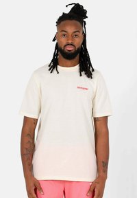 Crèmekleurig T-shirt met korte mouwen en een ronde halslijn, met een klein rood logo op de linker bovenborst. Zachte stoftextuur.