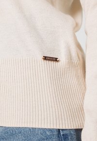 Jasnobeżowy dzianinowy sweter z prążkowanym wykończeniem na dole i przy mankietach. Z przodu, u dołu, wszyta mała, metalowa metka z logo "GUESS".