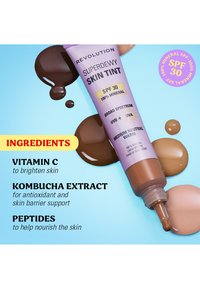 Tube de Superdewy Skin Tint avec SPF 30, teinte moyen neutre chaud, entouré d'échantillons de couleur et de la liste des ingrédients sur un fond bleu.