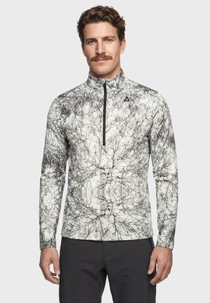 Mann mit Schnurrbart, der einen weißen und schwarzen Pullover mit abstraktem Muster und langen Ärmeln, einem schwarzen Reißverschluss und schwarzen Hosen vor einem schlichten Hintergrund trägt.
