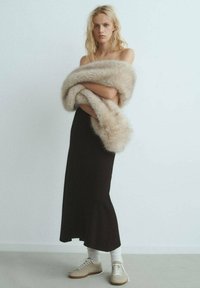 Stola in pelliccia sintetica color beige chiaro, indossata sopra un abito maxi aderente nero. Il modello indossa anche calze bianche e sneakers beige chiaro. Sfondo minimalista.