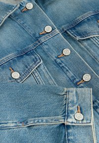 Veste en denim bleu clair avec boutons métalliques fermés, dotée de poches poitrine et de coutures orange détaillées.