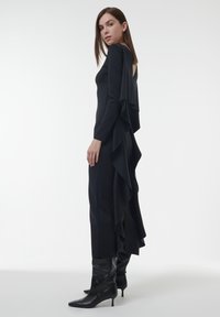 Leng zwarte jurk met lange mouwen, een diepe rug en een gelaagd ruchesdesign langs de achterkant. Gepaard met kniehoog zwarte laarzen.