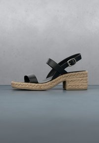 Sandale en cuir noir avec bout ouvert, bride à boucle et talon bas carré tissé texturé beige sur une surface grise.