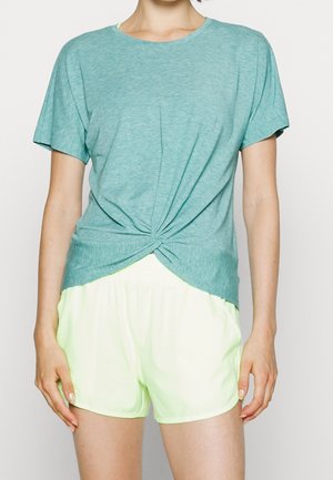 Woman wearing a teal short-sleeve knotted t-shirt and light yellow athletic shorts, standing against a plain background.