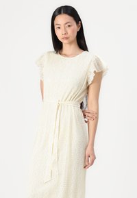 Robe ivoire arrivee au genou avec de courtes manches volantes, tissu texturé présentant des motifs géométriques, et une ceinture à la taille pour l'ajustement.