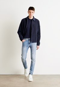 Camisa texturizada navy sobre un suéter de cuello redondo navy, combinada con pantalones jeans de corte slim de color azul claro y zapatillas blancas. Diseño minimalista, look casual.