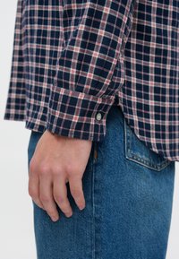 Tengerészkék kockás ing, piros és fehér vonalakkal, begombolt mandzsettával és puha anyaggal, kék farmernadrággal párosítva. Kézközelkép látható.