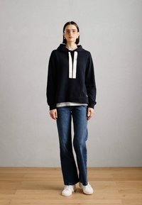 Juodas džemperis su baltais raišteliais, dėvimas ant pilkos palaidinės, derinamas su mėlynomis džinsinėmis kelnėmis ir baltomis sportbatėmis. Modelis stovi ant medinės grindų.