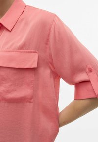 Camicia a maniche corte in tessuto corallo, con polsini abbottonati e tasca anteriore. Il materiale ha una texture liscia e leggera.