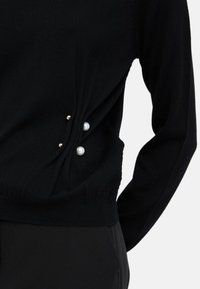 Maglione nero a maglia con dettagli arricciati sui lati, impreziosito da perle bianche e accenti dorati sul lato sinistro. Maniche lunghe su un corpo aderente.