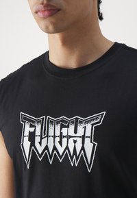 Muž ve bezrukávovém černém tričku s výrazným bílým grafickým textem "FLIGHT" v ostrém, stylizovaném písmu na hrudi na jednobarevném pozadí.