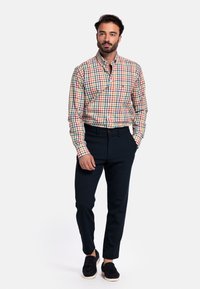 Geplooide button-up shirt in meerkleurige vierkanten, gecombineerd met strakke marineblauwe broek en zwarte instappers, op een model voor een neutrale achtergrond.