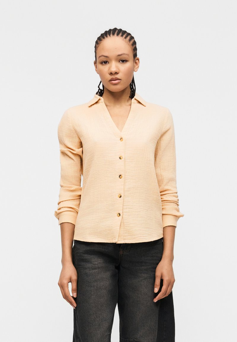 Jeune femme aux cheveux tressés portant une chemise à boutons couleur pêche clair et un jean foncé, debout devant un fond blanc uni.