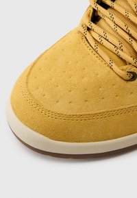 Dzeltens zamšādas sporta apavu ar teksturētu augšdaļu, tumši šņorēm un krēma krāsas gumijas zolīti. Apaļa pirksta daļa ar smalku šuvju detaļu.