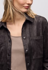 Camisa negra de ante sintético con cierre de botones, dos bolsillos en el pecho y un cuello clásico, llevada sobre una camiseta beige ajustada. Textura suave.