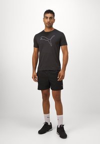 Tricou negru cu mânecă scurtă, cu logo alb Puma, purtat cu pantaloni scurți sport negri, șosete albe și adidași negri, stând pe un fundal neutru.