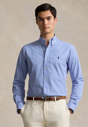 Kék-fehér csíkos, gombos ing klasszikus gallérral, hosszú ujjakkal és logó részlettel. Világos színű vászonnadrággal párosítva.