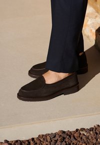 Brown suede slip-on shoes with a rounded toe and a stitched accent detail, paired with dark trousers on a light surface.