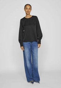 Blusa nera a maniche lunghe con scollo rotondo e polsini texturizzati, abbinata a jeans in denim blu a gamba larga e scarpe a punta.
