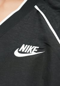 Fekete sportfelső fehér díszítésekkel, V-nyakú gallérral és hímzett Nike logóval. Az anyag sima, enyhén texturált felületű.