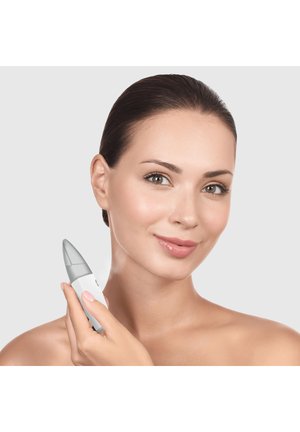 Femme à la peau claire tenant une brosse nettoyante pour le visage blanche et grise près de son cou, souriant doucement sur un fond clair uni.