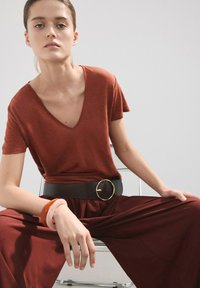 Jeune femme assise les jambes écartées, portant un haut à col en V de couleur rouille, un pantalon assorti, une large ceinture noire et des bracelets au poignet gauche.