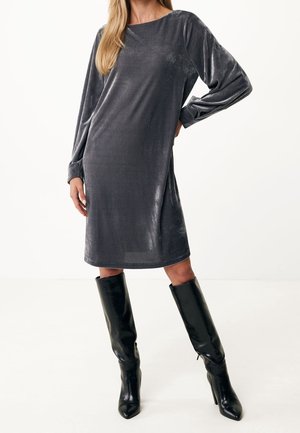 Frau trägt ein graues, knielanges Samtkleid mit langen Ärmeln, kombiniert mit schwarzen kniehohen Stiefeln mit Absatz, steht vor einem schlichten Hintergrund.