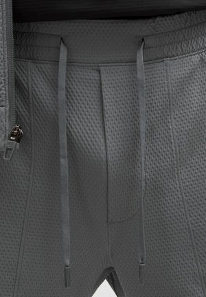 Primo piano di pantaloni grigio scuro con trama, coulisse e una cerniera parzialmente visibile sulla cintura del lato sinistro.