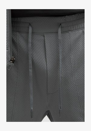 Detailní záběr tmavě šedých texturovaných kalhot s šňůrkami a částečně viditelným zipem na levé straně pasu.