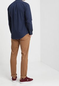 Man in een marineblauwe langemouwen shirt, beige broek opgerold bij de manchetten, en bordeauxrode sneakers, staand met handen in de zakken tegen een witte achtergrond.
