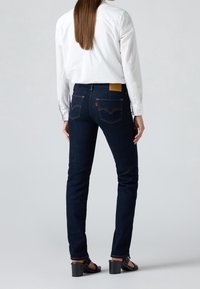 Personne portant un jean slim bleu foncé, une chemise blanche à manches longues et des sandales noires à talons, debout devant un fond clair uni.