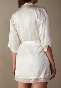 Sametový župan v jemné bílé barvě s páskem v pase. Obsahuje tříčtvrteční rukávy a krajkový lem na spodním okraji. Hladká textura a elegantní design.