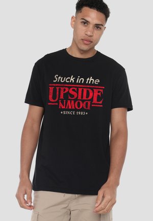 Young man wearing black T-shirt with red and white text "Stuck in the UPSIDE DOWN since 1983" standing against plain background.