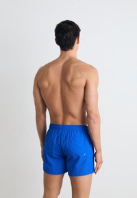 Un short de bain bleu avec une taille élastique et une poche au dos, fabriqué en matériau léger, vu de dos.