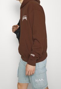 Hnědá mikina s kapucí a přední kapsou, na které je bílé text a "500" na manžetě. V kombinaci se světle modrými džínovými šortkami. Hedvábná textura.