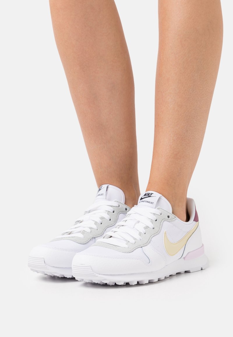 Personne portant des baskets blanches Nike avec un swoosh beige et des touches roses, debout devant un fond clair uni.