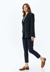 Marineblauwe getailleerde blazer met scherpe revers, lange mouwen en knoopsluiting, gecombineerd met slim-fit marineblauwe broeken en bruine leren instappers.